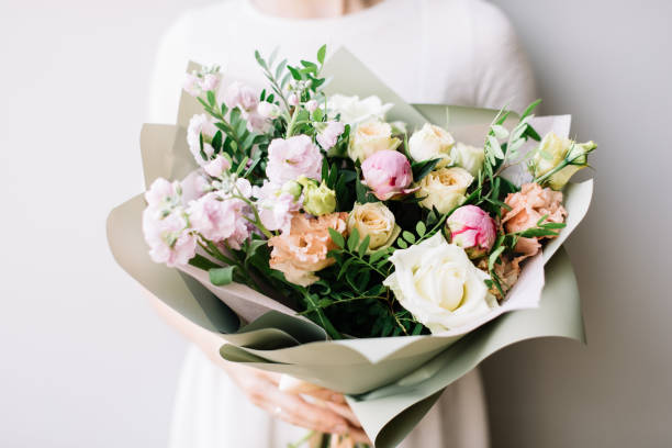 Very nice young woman holding beautiful blossoming bouquet of fresh roses, mattiola, eustoma flowers in pastel pink and cream colors on the grey wall background stock photo