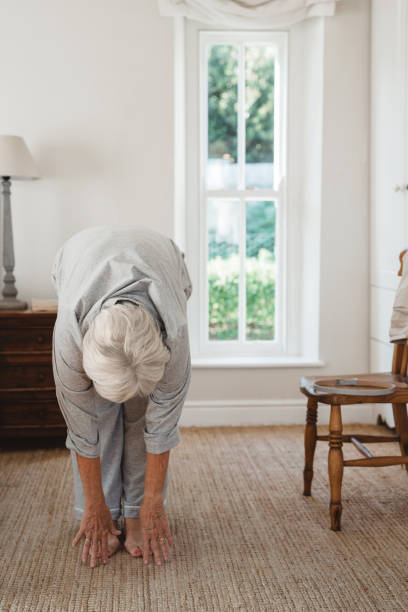 Senior woman doing stretches after waking up in the morning Senior woman in pajamas stretching to touch her toes while waking up in her bedroom in the morning touching toes stock pictures, royalty-free photos & images
