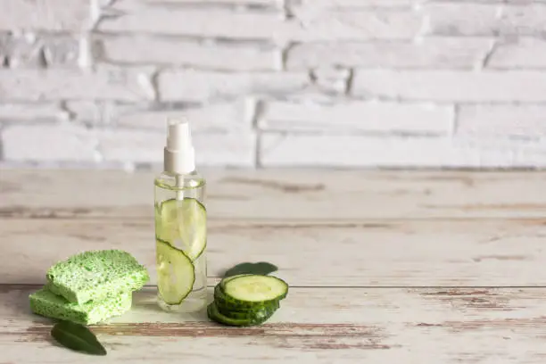 Homemade refreshing facial cucumber toner in bottle with slices of green cucumber and cotton-pads on white wooden background. Copy space.