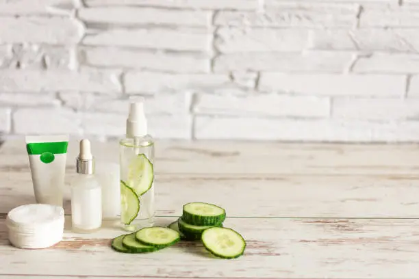 Bottles of homemade cucumber natural cosmetic with green cucumber slices and cotton-pads on wooden background. Copy space.
