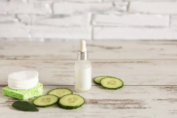 Bottle of homemade facial cucumber organic serum with green cucumber slices and cotton-pads on wooden background.Copy space.