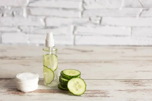 Homemade refreshing facial cucumber toner in bottle with slices of green cucumber and cotton-pads on white wooden background. Copy space.