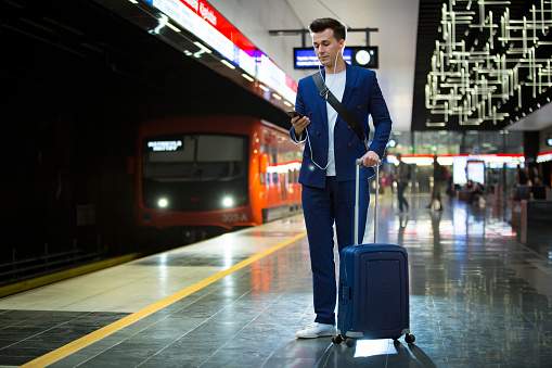 Young stylish handsome man in suit with suitcase standing on metro station holding smart phone in hand, scrolling and texting, smiling and laughing on subway station. Finland