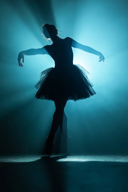 istock Young beautiful ballerina on smoke stage dancing modern ballet. Performs smooth movements with hands against spotlight background. Woman in black tutu costume on scene. 1188900772