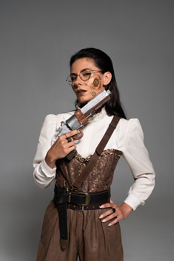 attractive steampunk woman standing with hand on hip and holding pistol isolated on grey