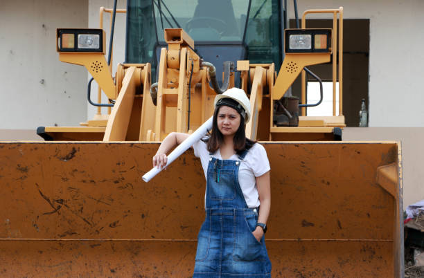 istock Female civil engineer or architect with white helmet and project drafts while in hand standing with Alloy Bulldozer Truck with Front Loader Truck Engineering Construction Car Vehicle. 1188907097