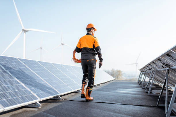 Well-equipped workman on a solar station Well-equipped worker in protective orange clothing walking and examining solar panels on a photovoltaic rooftop plant. Concept of maintenance and installation of solar stations ecosystem photos stock pictures, royalty-free photos & images