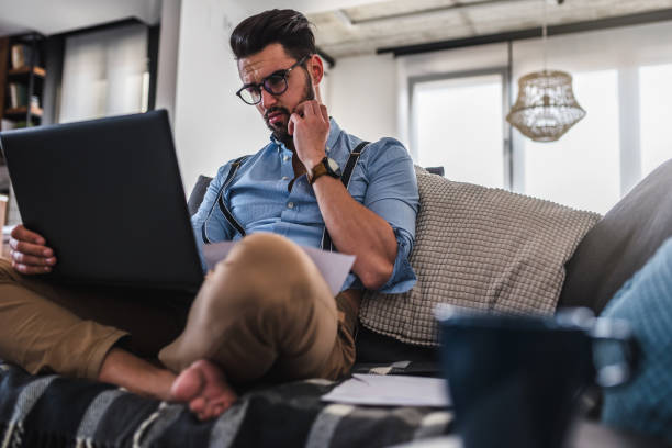 istock Young bearded businessman working on laptop computer while sitting on sofa at his home office. Business problems. 1188911925