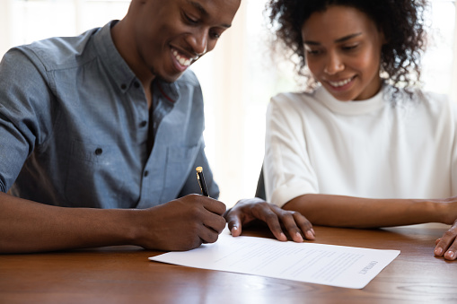 Focus on male hands, holding pen, signing contract. Happy african ethnicity young woman watching smiling husband putting signature on agreement. Glad mixed race family couple buying property.