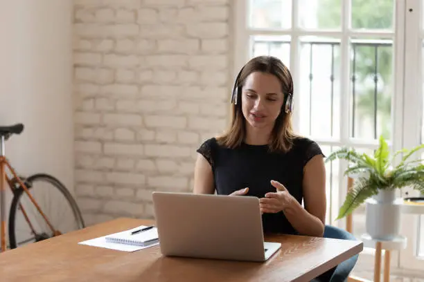 Confident young businesswoman wearing wireless headphones, discussing project details with clients via video call. Intelligent skilled female worker speaker holding online meeting with colleagues.