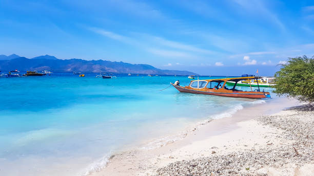 Gili Island - A boat anchored to a white sand beach A boat parked next to the shore on Gili Air, Lombok, Indonesia. Beautiful and clear water. In the back visible Mount Rinjani. Some trees on the shore, few clouds on the sky. Holidays paradise. gili trawangan stock pictures, royalty-free photos & images
