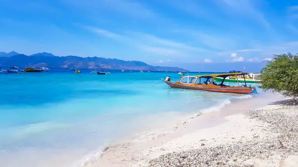 A boat parked next to the shore on Gili Air, Lombok, Indonesia. Beautiful and clear water. In the back visible Mount Rinjani. Some trees on the shore, few clouds on the sky. Holidays paradise.