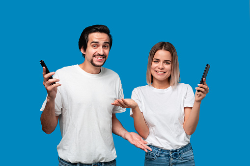 Blond woman and brunet man in white tees and blue jeans are surprised and shaking shoulders holding smartphones and looking at the camera. Good news.