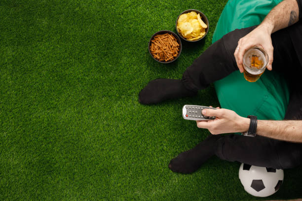 A man sits at the TV and watches a football match. Sneki TV remote. Soccer ball. View from above. Copy space. Imitation of a lawn. stock photo