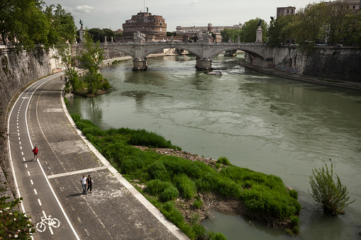 Horizontal high angle view of some people strolling and running along Tiber River with Ponte Vittorio Emmanuelle II and the Castel Sant’ Angelo in the background, Rome