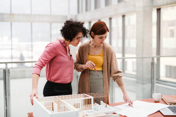 weibliche junge architekten mit modell eines hauses im büro stehen, reden. - architekturberuf stock-fotos und bilder