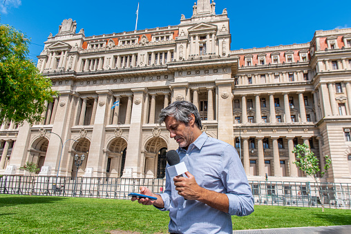 Male mature reporter talking, holding a microphone and looking at his mobile phone in a public park during a sunny day at Buenos Aires, Argentina. At the background the facade of the Palace of Justice.