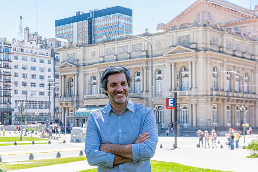 Portrait of a mature man with his arms crossed smiling in a public park during a sunny day at Buenos Aires, Argentina. At the background the facade of the Colon Theater.