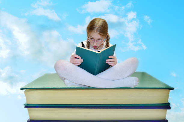 una studentessa in uniforme scolastica si siede a gambe incrociate su grandi libri e legge un libro a sorpresa. contro il cielo blu con nuvole bianche. il concetto di apice della conoscenza, l'interesse per l'apprendimento. - childrens literature foto e immagini stock