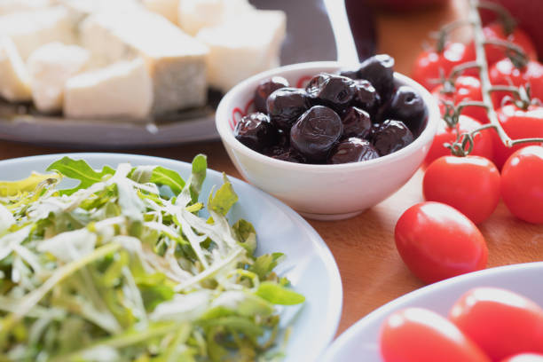 Black olives in small bowl on table, with arugula leaves, soft cheese and cherry tomato, close up
