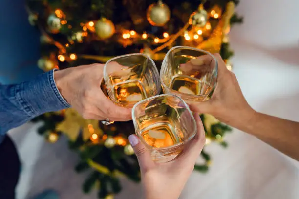 Close up on three female women hands holding a glasses of whiskey or brandy or cognac alcohol drink toasting celebrating at home by the christmas tree