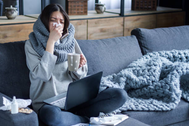 Sick female with cold and flu. Health care concept Ill brunette girl is sitting at sofa and working remotely on laptop. Female is blowing out snot, having fever and headache. Young woman is treated at home, took sick leave. Winter cold and flu concept flu virus stock pictures, royalty-free photos & images