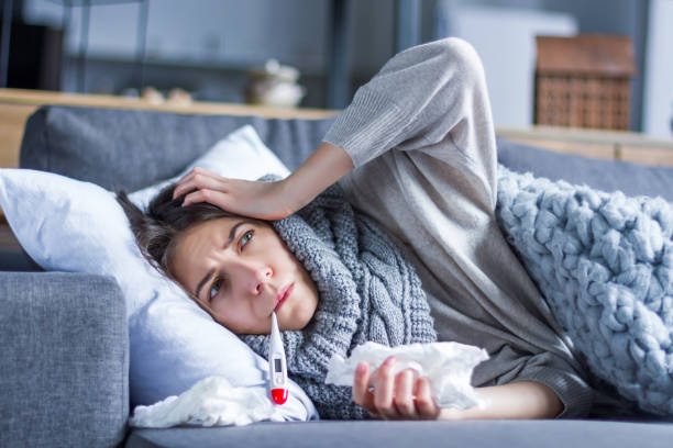 Sick female with cold and flu. Health care concept Sick exhausted girl in scarf is lying in bed wrapped in blanket. Young woman with fever and headache is measuring temperature with thermometer, treated at home. Winter cold and flu concept. flu virus stock pictures, royalty-free photos & images