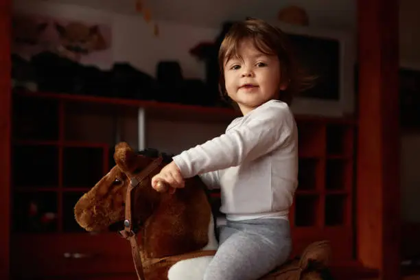 Little baby girl sitting on rocking horse, playing, being indoors and feeling happy, looking at camera