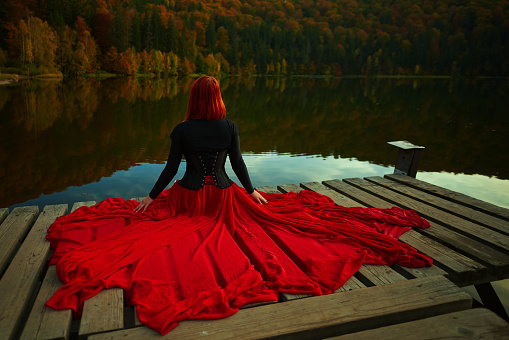 Rear view of woman wearing red skirt posing in fairytale concept at the lake, autumn concept, sitting on wooden pontoon and thinking