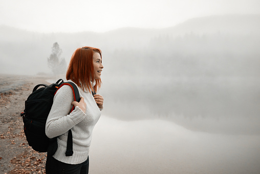 Profile view of happy redhead backpacker standing on the edge of the lake, smiling and admiring the fog