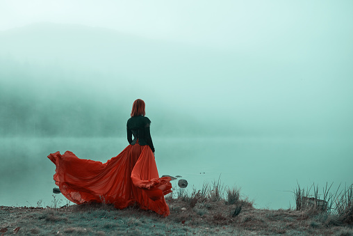 Rear view of woman wearing red skirt posing in fairytale concept at the lake, autumn concept, fog surrounding her