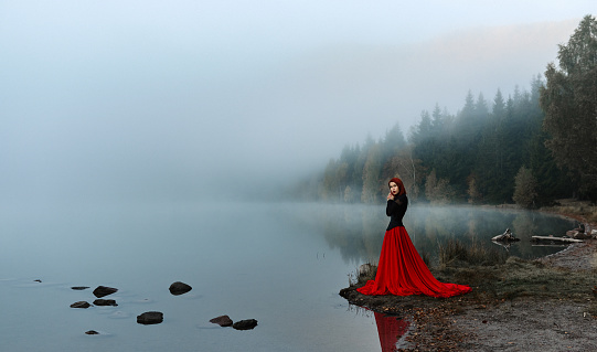 Woman wearing red skirt posing in fairytale concept at the lake, autumn concept, fog surrounding her