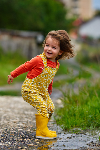 Little baby girl wearing yellow rubber boots playing outside in the mud on a rainy day, jumping and splashing