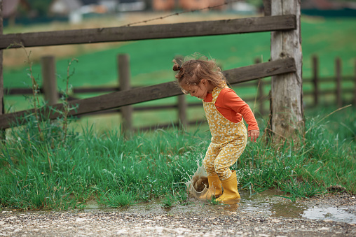 Little baby girl wearing yellow rubber boots playing outside in the mud on a rainy day, splashing and jumping