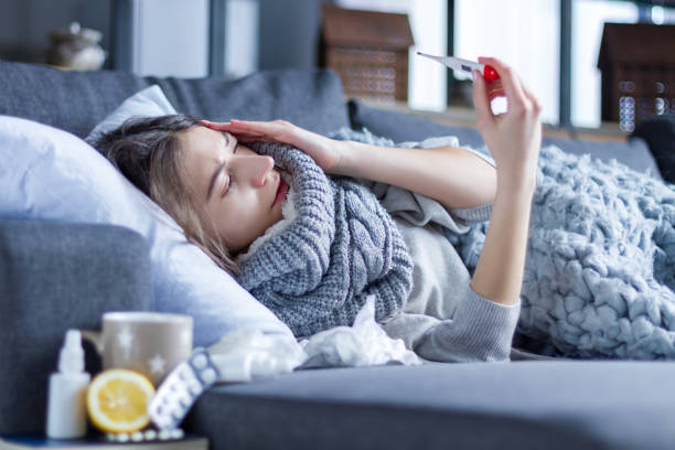 Sick female with cold and flu. Health care concept Sick exhausted girl in scarf is lying in bed wrapped in blanket. Young woman with fever and headache is measuring temperature with thermometer, treated at home. Winter cold and flu concept. cold and flu stock pictures, royalty-free photos & images