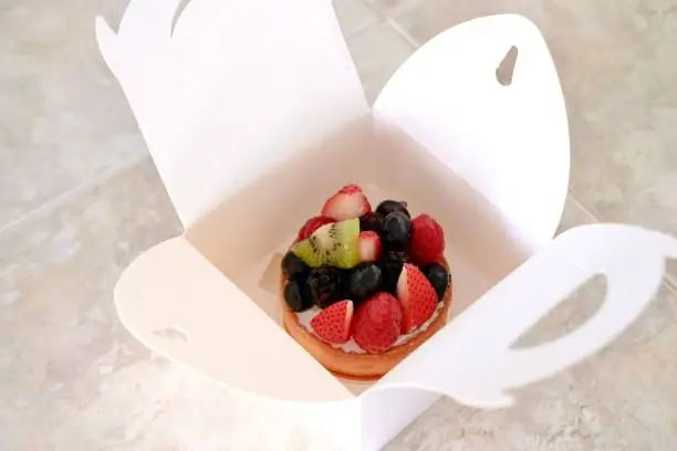 Photo of Fresh fruit tart in white paper box on table. Modern pastry concept.