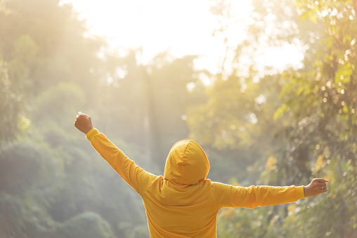 Happy woman traveler in yellow sweater with hood is standing with raised arms over nature on vacation concept. Rear view.