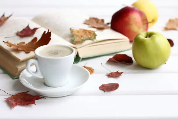 white cup with coffee on the background of blurry outlines of an open book and apples with autumn leaves