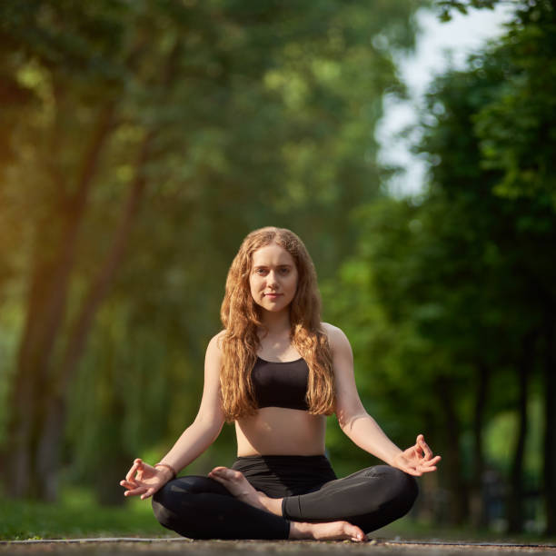 Cute girl is meditating in lotus position, Padmasana Asana on the background of blurred green park Cute girl meditating in the lotus position, Padmasana Asana on the background of a blurred green park under the sun's rays on a warm day. Morning yoga training outdoor all day energy boost religion sunbeam one person children only stock pictures, royalty-free photos & images
