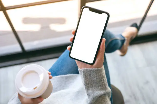 Photo of a woman holding a black mobile phone with blank white desktop screen with coffee cup