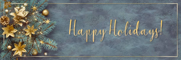 Gilded text Happy Holidays. Christmas or New Year panoramic background - fir twigs decorated with golden trinkets, flowers and Xmas lights. Top view on dark background with shiny golden frame.. stock photo
