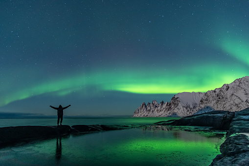 Man watching the northern lights, Aurora Borealis, Devil Teeth mountains in the background and a rock formation and the ocean in the foreground, Tungeneset, Senja Island, Troms, Norway, Europe