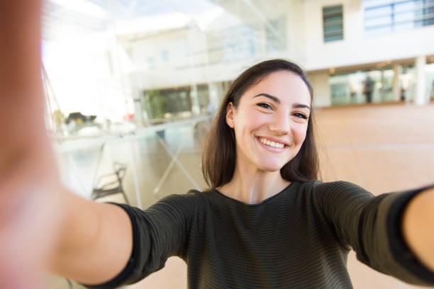 Self portrait of joyful beautiful woman holding smartphone Self portrait of joyful beautiful woman holding smartphone with both hands. Young woman in casual posing indoors with glass wall interior in background. Taking selfie concept women taking selfies photos stock pictures, royalty-free photos & images