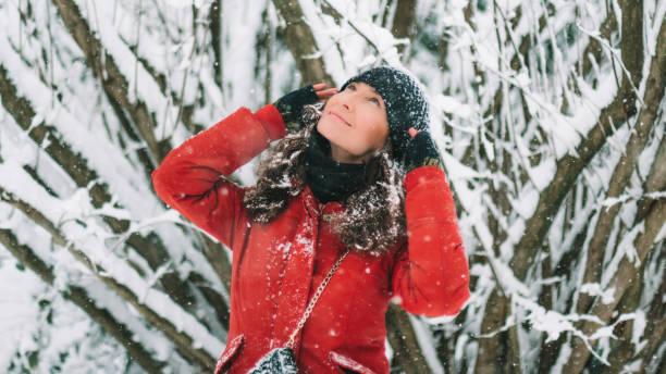 istock Young woman in red down jacket and black hat looking up at snow flakes while outside is snowing 1189097516