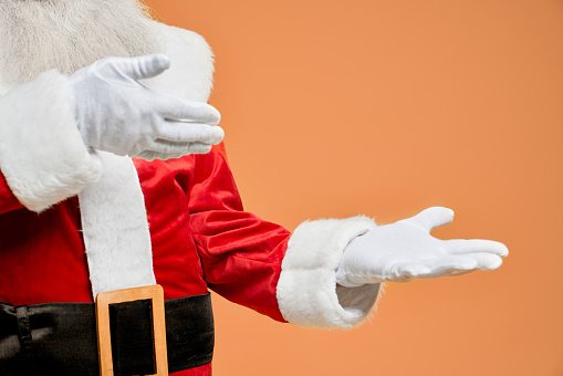 Close up of Santa Claus hands in white gloves with open palms and empty space posing in studio with orange background. Place for text or advertisement of some product.