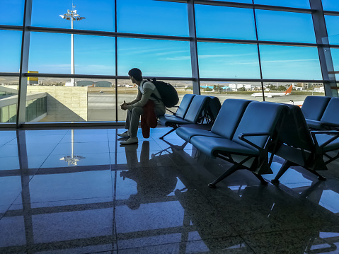 Passenger sits in the departure zone of the airport and looks at the runway through the panoramic window. Male tourist with backpack waiting for his plane, concept of cancellation or delayed flight