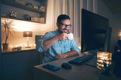 Handsome smiling freelancer working remotely from home and drinking coffee. He is working at night.
