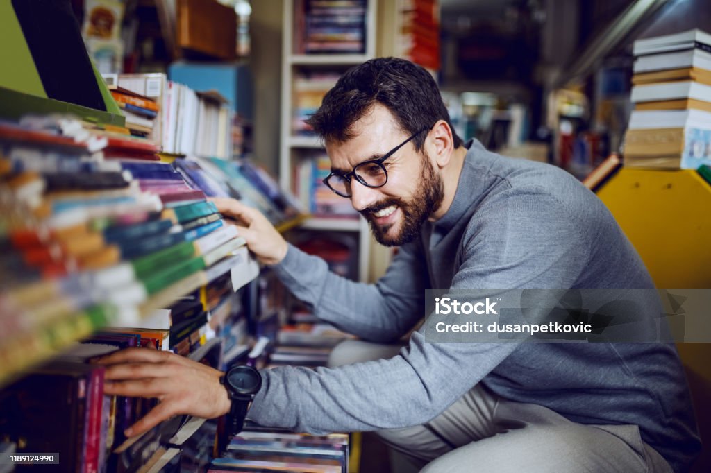 Young handsome caucasian bearded man with eyeglasses crouching in bookstore and searching for interesting book. Bookstore Stock Photo