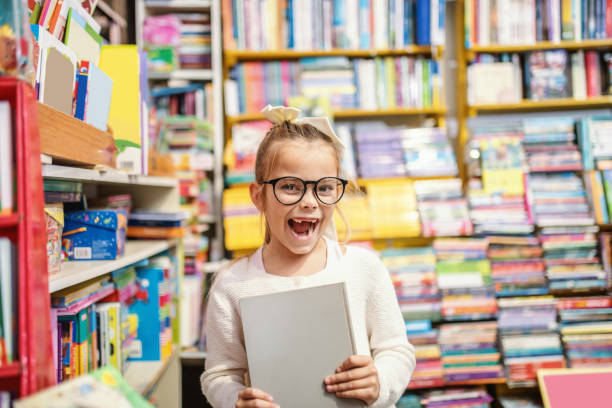 animado caucasiano menina loira bonito com óculos e rabo de cavalo de pé na livraria e segurando livro que ela estava procurando. - child glasses elementary student reading - fotografias e filmes do acervo