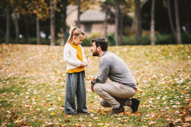 istock Devilish cute little girl with ponytail standing and looking at her father. Father crouching and reprimanding her. Autumn in park. 1189128184